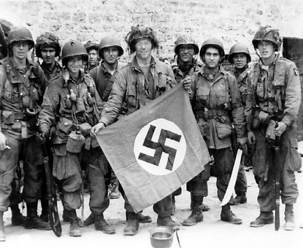 American paratrooper James Flanagan (2nd Platoon, C Co, 1-502nd PIR), among the first to make successful landings on the continent, holds a Nazi flag captured in a village assault. France. 6 June 1944